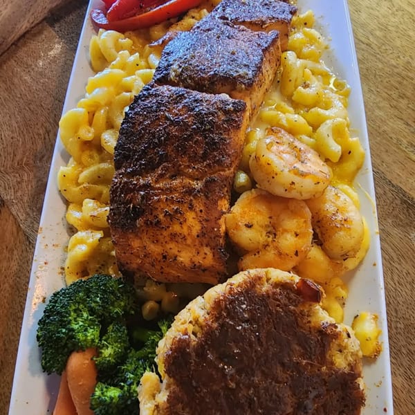 Grilled salmon, shrimp, crab cake, macaroni and cheese, broccoli, and carrots on a white plate.